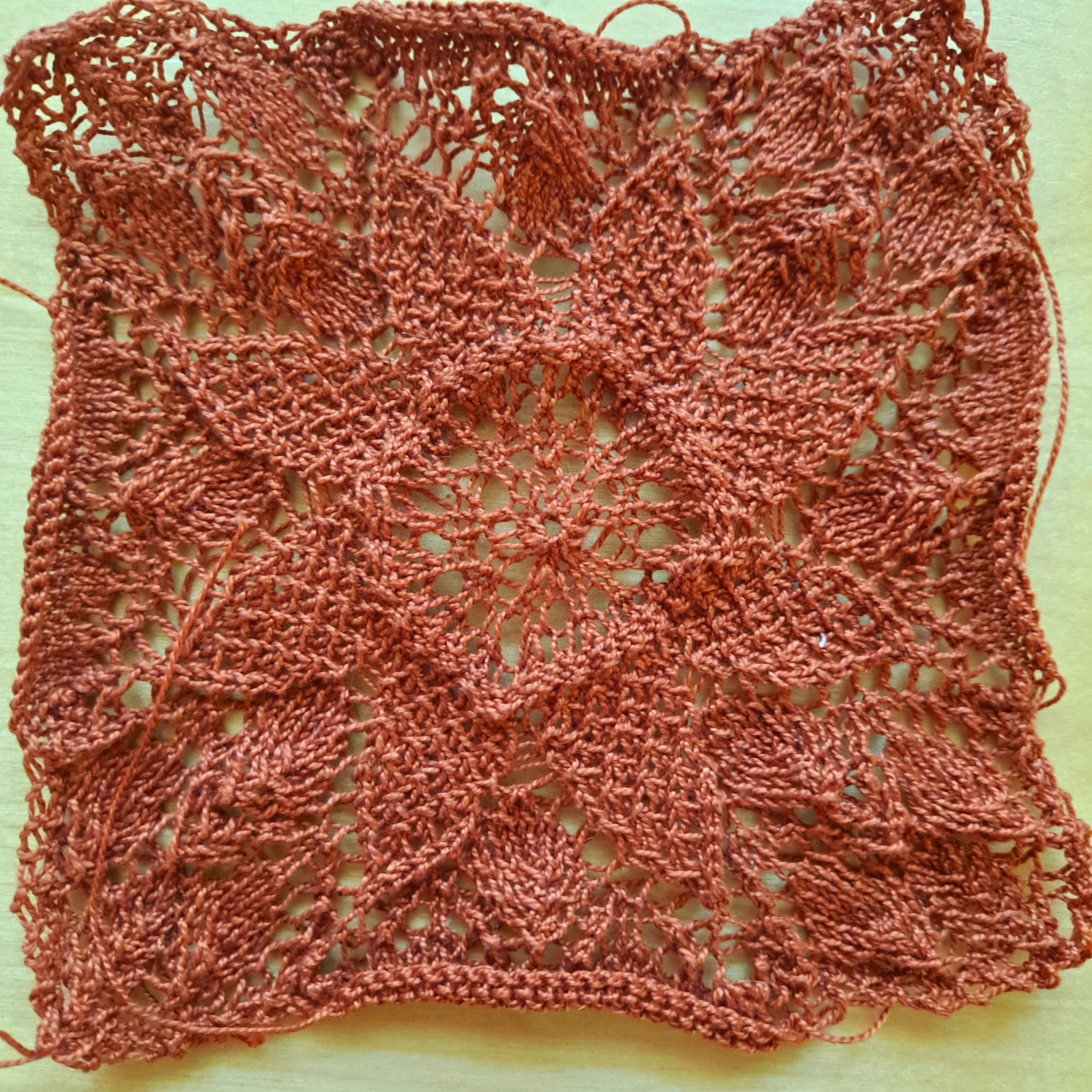 An image of a lacy square knitted in copper yarn with an eight-petaled flower worked into the lace